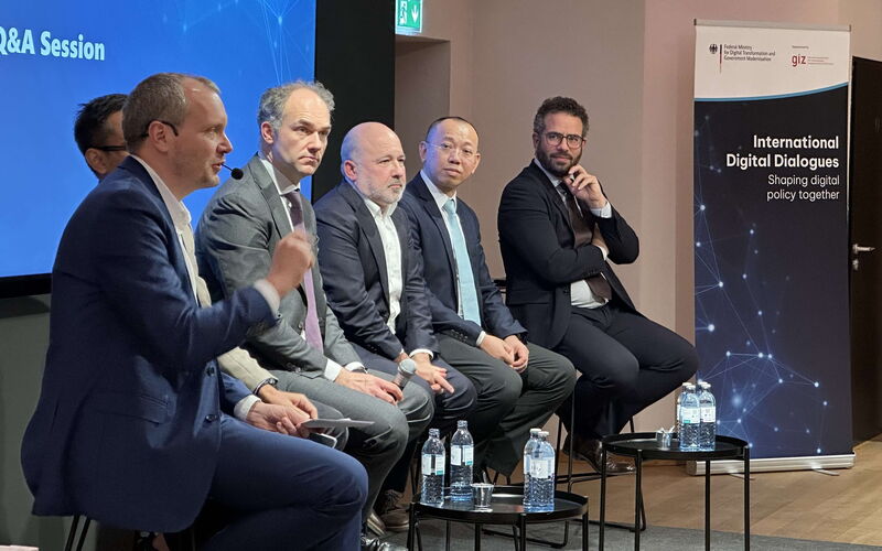 Five panelists are sitting on the stage looking right to the moderator sitting at the right end doing the introduction to the paneldiscussion