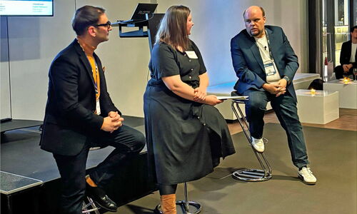 Three people are seated on bar stools on a stage during a panel discussion. Two men and one woman are engaged in conversation. Behind them, a screen displays a presentation, and a lectern stands nearby. The room has a modern setting with dim lighting.