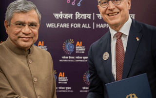 Zwei Männer in formeller Kleidung schütteln sich die Hand bei einem offiziellen Treffen vor einer Wand mit der Aufschrift ‚AI Impact Summit‘