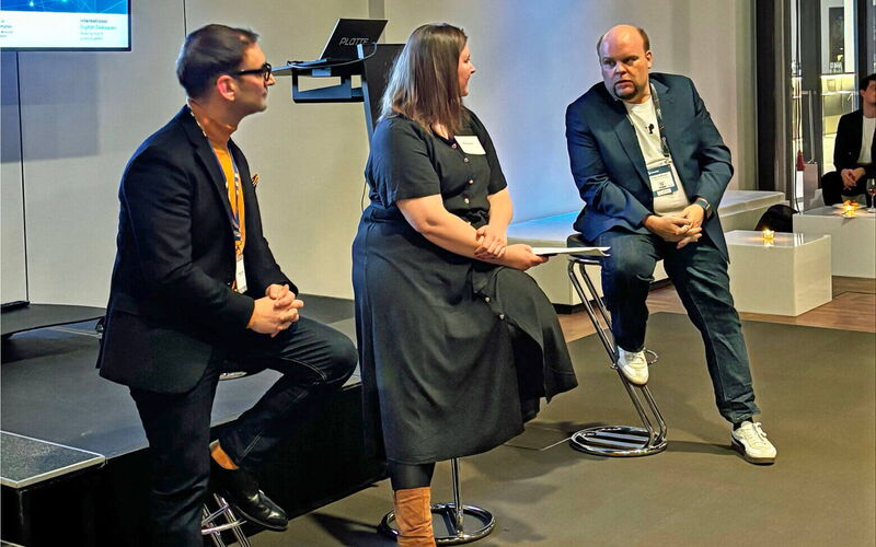 Three people are seated on bar stools on a stage during a panel discussion. Two men and one woman are engaged in conversation. Behind them, a screen displays a presentation, and a lectern stands nearby. The room has a modern setting with dim lighting.
