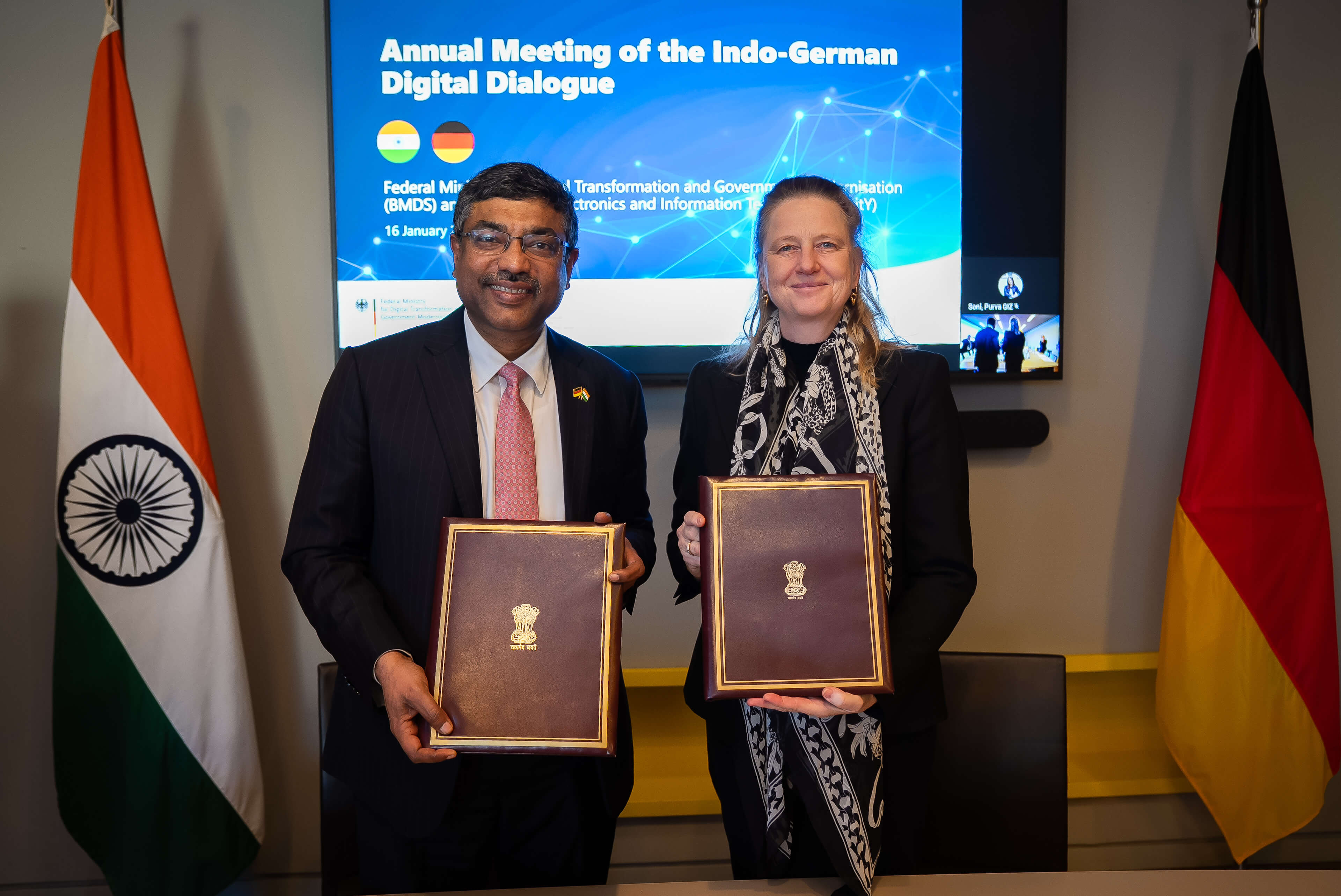 Zwei Personen stehen vor einer Präsentationswand zwischen der indischen und der deutschen Flagge und halten jeweils eine unterzeichnete Mappe in den Händen, was auf eine formelle Vereinbarung im Rahmen des deutsch-indischen Digitaldialogs hinweist.