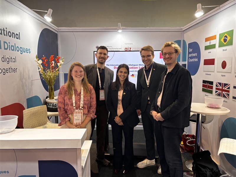 Fünf Personen stehen gemeinsam lächelnd am Stand der International Digital Dialogues beim India AI Impact Summit, umgeben von Informationsmaterialien und Flaggen der Partnerländer.